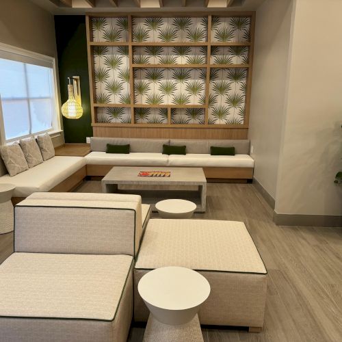 A modern lounge area with beige seating, a coffee table, tropical-patterned wall, potted plant, and green cushions in a well-lit room.