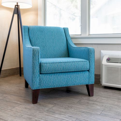 A blue armchair is placed near a window beside a floor lamp and a white air conditioner in a bright, modern room with wooden flooring.