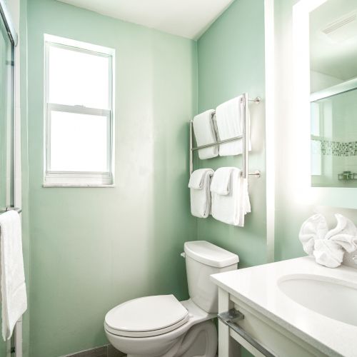 A clean bathroom with a shower, toilet, and sink. Towels are neatly hung, and the room is brightly lit by a window and mirror light.