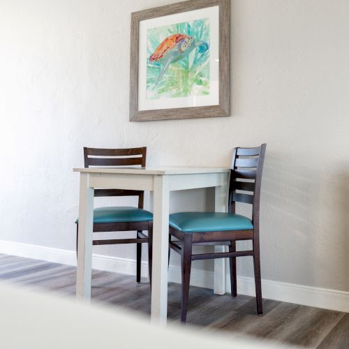 A small dining table with two chairs, a colorful framed fish artwork on the wall, and a bright, minimalist room design with wood flooring.