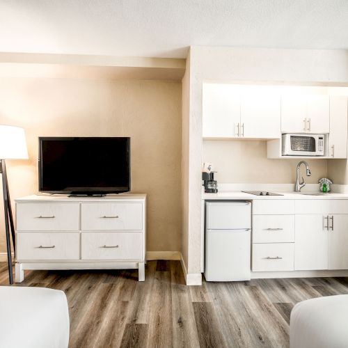 The image shows a hotel room with a TV on a dresser, a kitchenette with white cabinets, a sink, a microwave, and a refrigerator.