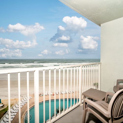 An ocean view from a balcony with two chairs, overlooking a beach and a pool, under a blue sky with scattered clouds.