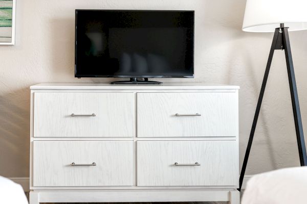 A small flat-screen TV is placed on top of a white dresser, which has four drawers, next to a floor lamp with a tripod base.