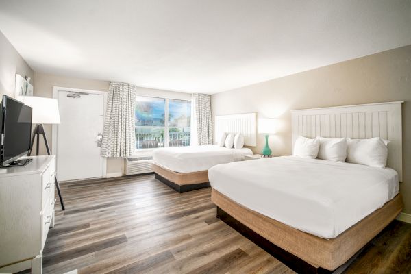 A clean, bright hotel room with two double beds, wooden flooring, a flat-screen TV, a window with curtains, and bedside tables with lamps.