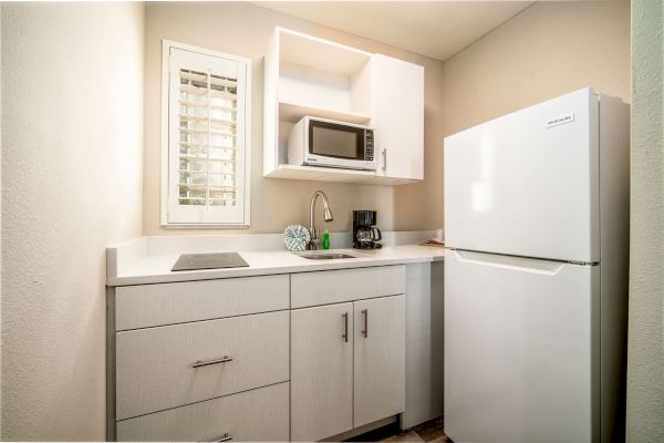 A compact kitchen with a fridge, microwave, sink, coffee maker, and cabinets, all in a clean, modern design with white and light wood tones.