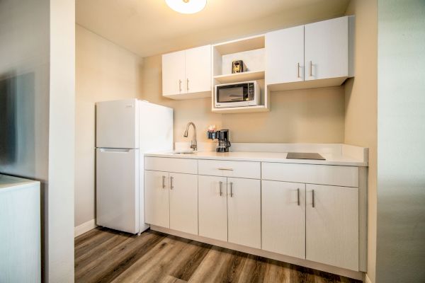 This image shows a compact modern kitchenette with white cabinets, a refrigerator, microwave, sink, and coffee maker on wooden flooring.