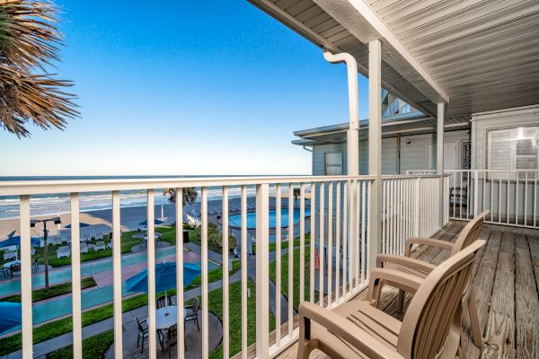 A balcony with two wooden chairs overlooks a beachfront, with views of a pool, beach umbrellas, and the ocean under a clear blue sky.