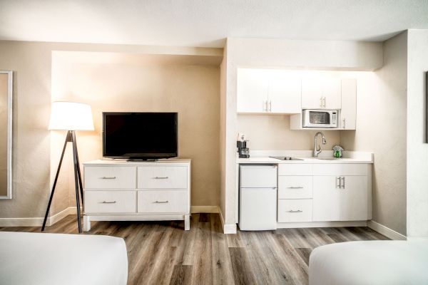 The image shows a hotel room with a flat-screen TV on a dresser, a kitchenette with white cabinets, a small fridge, a microwave, and a lamp.
