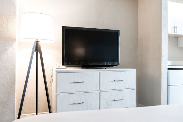 A black TV on a white dresser with four drawers next to a floor lamp with a white shade in a bright room.