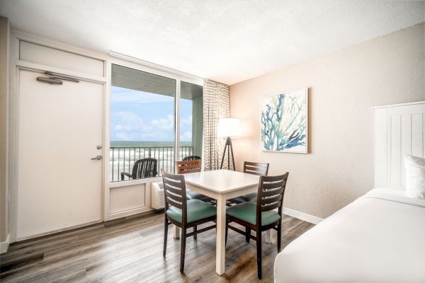 The image shows a bright room with a table and four chairs, a bed, a floor lamp, wall art, and a door leading to a balcony with an ocean view.