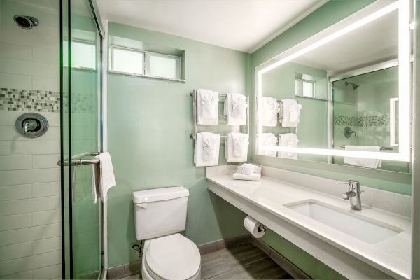 A modern bathroom with a glass shower, toilet, and large illuminated mirror above a sink counter; towels are neatly hung on a rack.