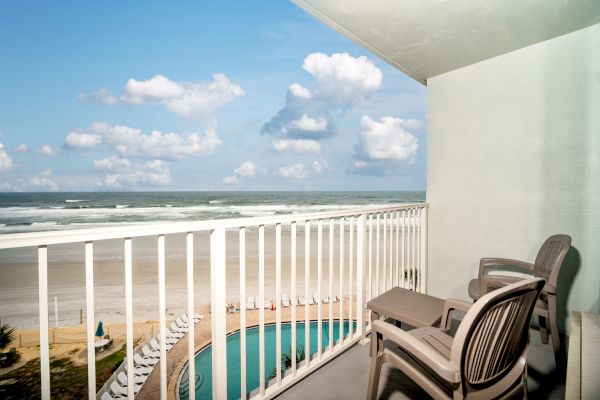 A balcony overlooking a beach, featuring a pool, two chairs, a small table, and the ocean under a blue sky with scattered clouds.