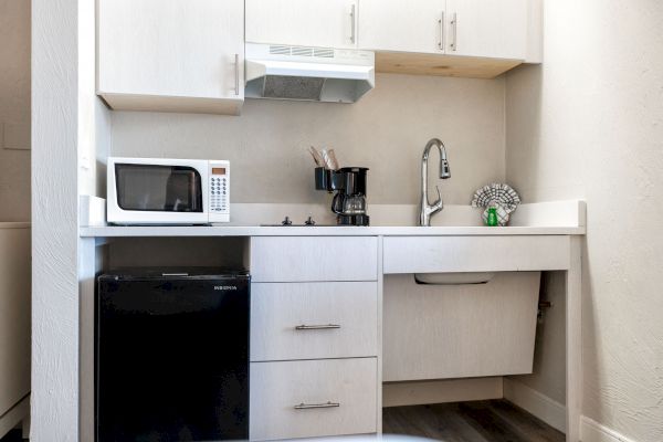 A compact kitchenette with a microwave, coffee maker, sink, faucet, small fridge, and cabinets above, all in a light-colored finish.