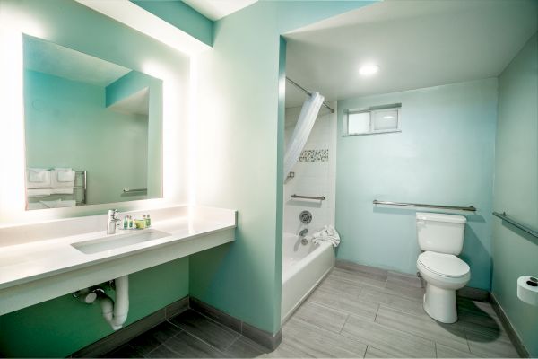 A modern bathroom with a large mirror, sink, toilet, bathtub with shower curtain, and teal walls. The room is well-lit with a minimalist design.
