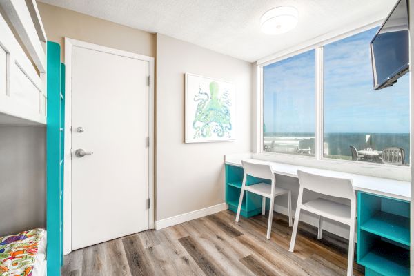 The image shows a bright room with a door, artwork of an octopus, windows with an ocean view, a desk with two chairs, and a wall-mounted TV.