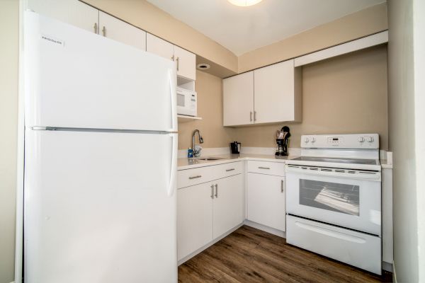 A small kitchen with white cabinets, a refrigerator, microwave, sink, countertop appliances, and a stove with an oven, all on wood flooring.