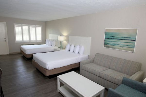 The image shows a hotel room with two beds, a sofa, an armchair, and a coffee table, featuring a serene beach painting on the wall.