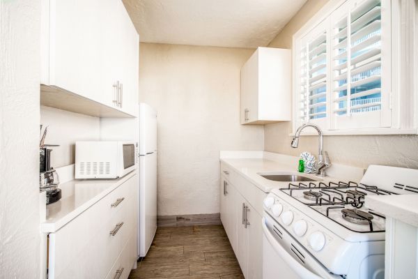 A small, bright kitchen with white cabinets, a gas stove, microwave, refrigerator, and a sink under a window with blinds.