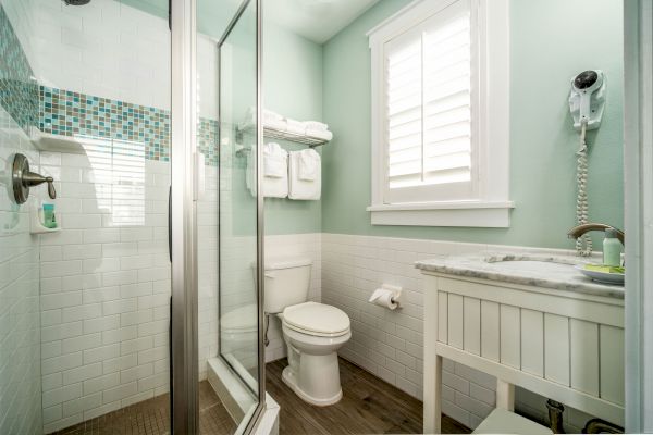 This image shows a modern bathroom with a glass shower, toilet, sink with countertop, wall-mounted hairdryer, and towels on a shelf, all in a pastel green.