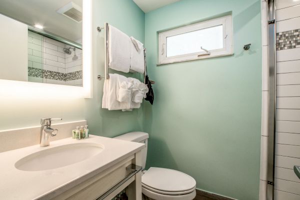 This image shows a modern bathroom with a light green wall, a sink with a mirror, a towel shelf, a toilet, and a shower area on the right.