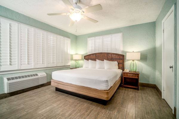 A modern bedroom with a large bed, two glass bedside lamps, a ceiling fan, green walls, wooden flooring, and white shutters on the windows ends the sentence.