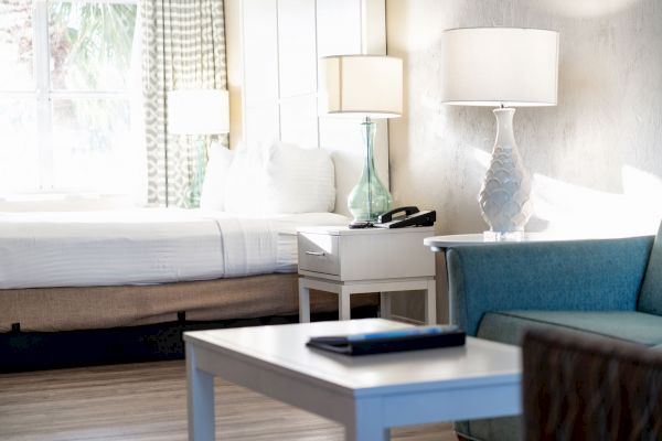 A well-lit hotel room with a bed, nightstands, lamps, a couch, and a coffee table with a closed tablet on it.