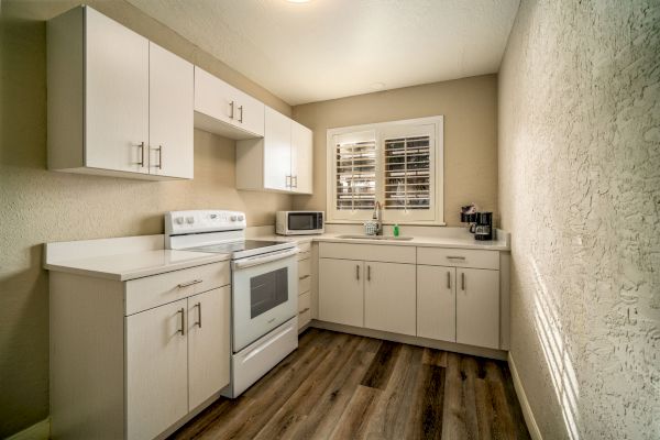 A small, modern kitchen with white cabinets, a stove, microwave, and coffee maker. The room has wooden flooring and a window with blinds.