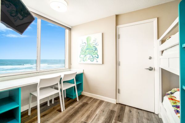 A room with a beach view, featuring a desk with two chairs, a bunk bed, and an octopus painting on the wall, all in a coastal-themed decor.