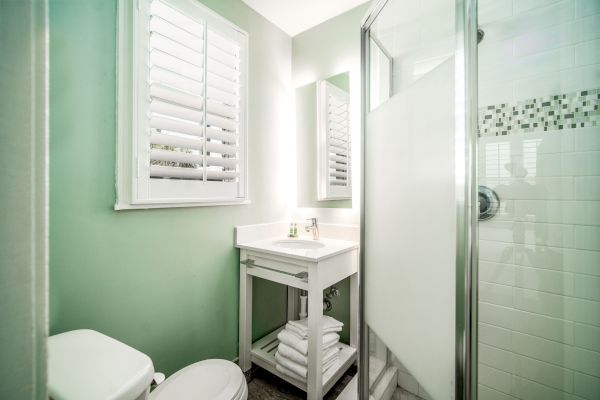 A small, modern bathroom with a shower, sink, white towels, toilet, and a window with shutters in a light-green painted room ending the sentence.